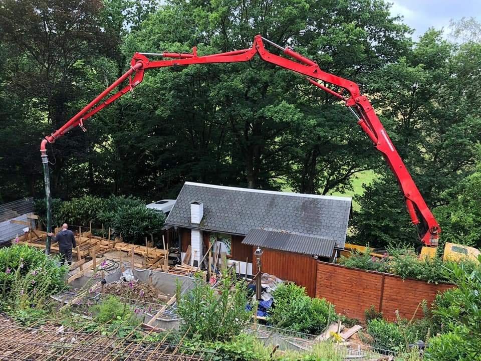 Bauarbeiten an einem kleinen Gartenhaus, während ein roter Betonmischer die Betonschicht aufträgt, um das Fundament zu gießen, in einem Garten mit Bäumen und Grünflächen.