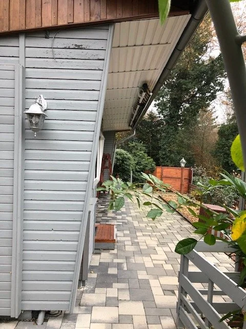 Hinterhof mit gepflastertem Weg, Zäunen und Bäumen, seitlicher Blick auf eine Hauswand mit Laterne.