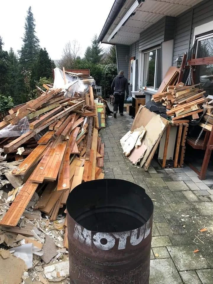 Ein Haus mit einer Terrasse, auf der Holzreste, Bretter und Baumaterialien gestapelt sind. Im Vordergrund steht ein leerer, bemalter Metallcontainer. Zwei Personen sind im Hintergrund unterwegs, eine trägt eine dunkle Jacke.