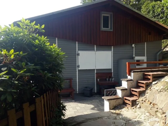 Ein Haus mit grauen Wänden, rotem Dach und kleinen Fenstern, umgeben von Bäumen, mit einer Treppe aus Ziegeln und Holz, im Freien.
