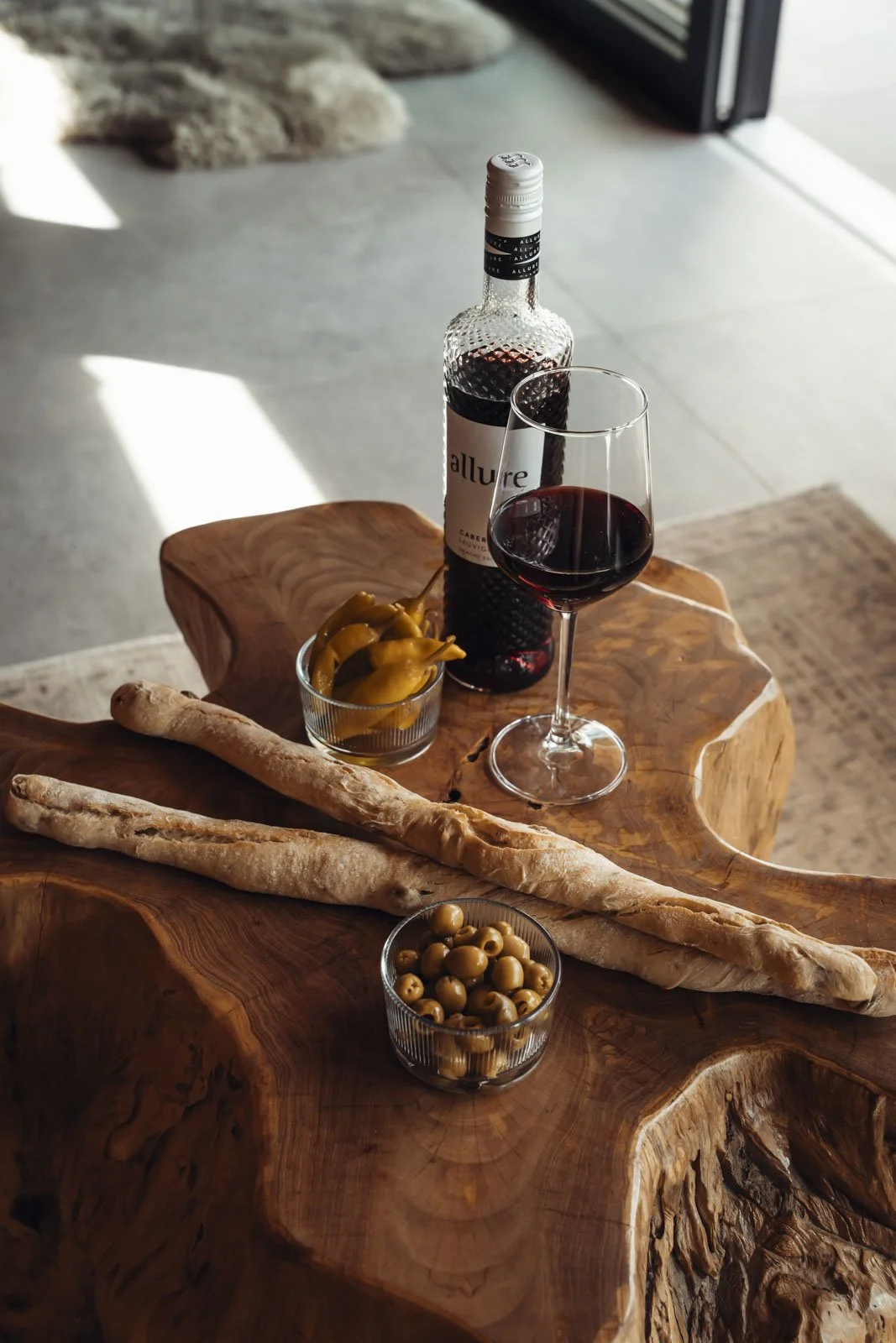 Ein Holztablett mit einer Flasche roten Weins, einem Weinglas, zwei Baguettes, grünen Oliven in einer Schüssel und eingelegte Paprika, im Hintergrund ein Teppich auf einem Boden