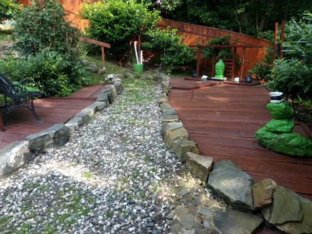 Gartenweg mit Kies und Steinen, umgeben von Holzdecks, grünem Laub und Skulpturen
