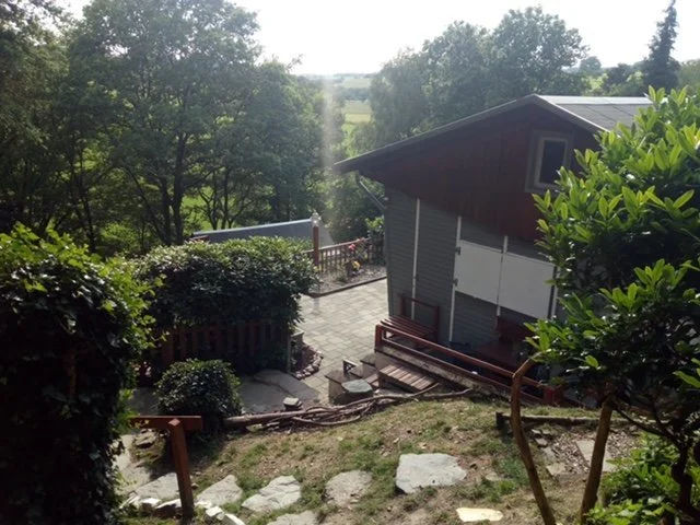 Gartenblick auf ein Haus mit Terrasse, umgeben von Bäumen und Büschen, mit Sonnenlicht.