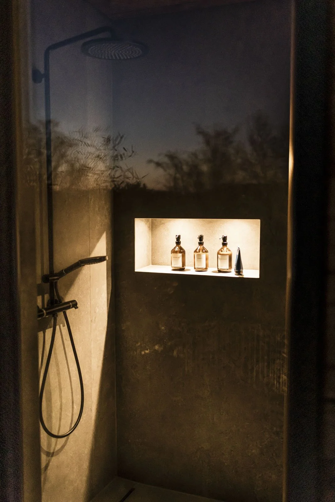 Duschkabine mit einer Handbrause auf der linken Seite und drei Flaschen auf einer Nische in der Wand, beleuchtet mit warmem Licht, im Hintergrund ist eine Fensterfront, durch die Bäume bei Sonnenuntergang zu sehen sind.