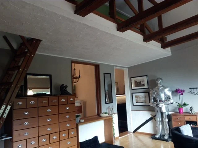 Innenansicht eines Wohnzimmers mit Treppe, Kommode, Spiegel, Bildern, einem Deko-Helm und einer Rüstung, Pflanzen und Möbeln.