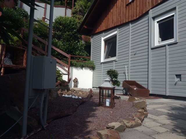 Gartenweg neben einem Haus mit grauer Fassade, zwei Fenster, ein Weg aus Pflastersteinen, ein kleiner Baum, eine Laterne, ein rotes Blumentrog, Steine und eine Kabelbox