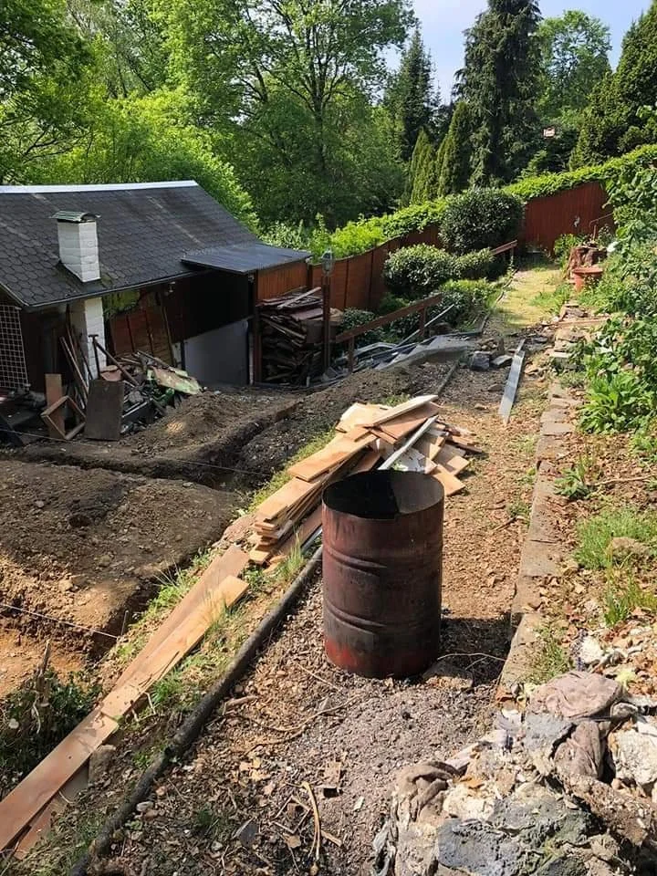 Garten im Umbau mit Bauholz, einem alten Metallfass und einem Graben, umgeben von Bäumen und grünen Pflanzen.