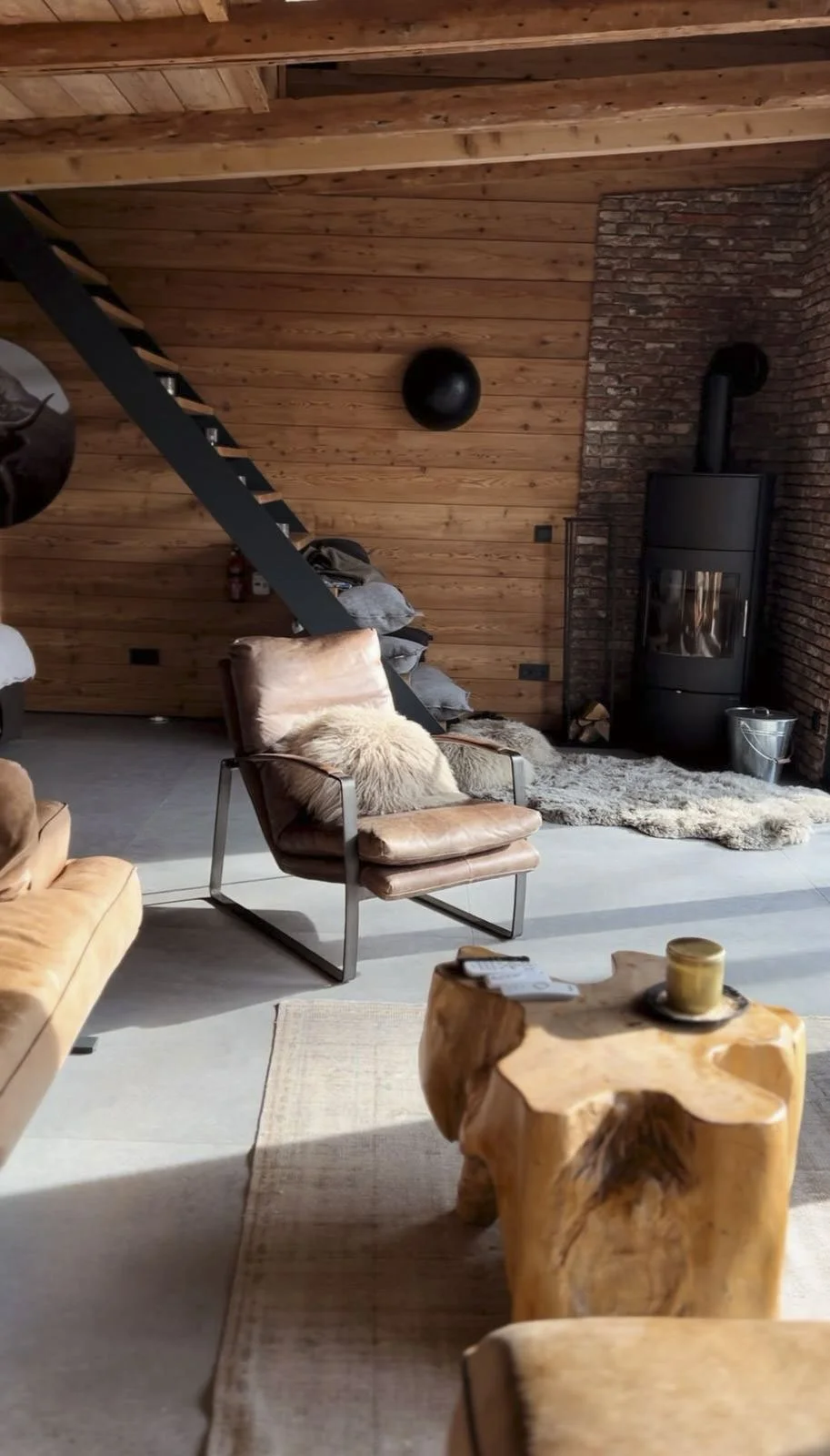Gemütliches Wohnzimmer mit Holzdecke und -Wänden, einem schwarzen Kamin, geflochtenen Teppichen, einer Lederliege mit Fellkissen und einem rustikalen Holztisch.