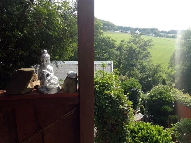 Ein Blick von einer Veranda auf eine grüne Landschaft mit Bäumen, Wiesen und Hügeln im Hintergrund. Auf der Veranda stehen silberne Buddha-Figuren und Steine.