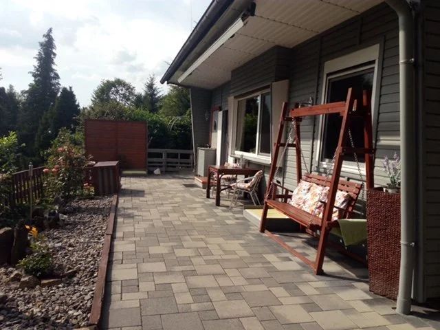 Blick auf eine Terrasse mit Holzmöbeln, einem Schaukelstuhl, einem Blumentopf und Gartenstühlen vor einem Haus mit Vinylverkleidung.