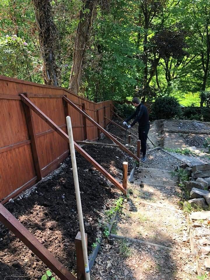 Eine Person arbeitet im Garten, befestigt einen Holzzaun entlang eines Weges, umgeben von Bäumen und grüner Natur.