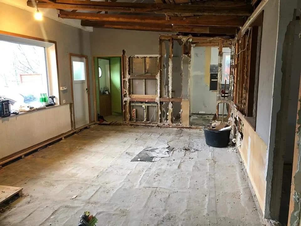 Innenaufnahme eines Raums, in dem Wände entfernt wurden, um eine Renovierung durchzuführen. Es ist Bauordnung sichtbar, mit lockerem Bodenbelag, einem großen Fenster auf der linken Seite und einem Durchgang in einen kleinen Raum. Baustoffreste und We