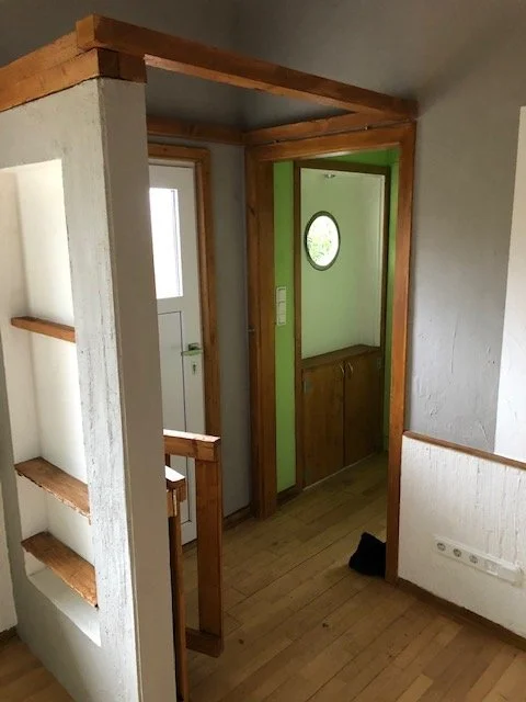  Blick in einen Eingangsbereich mit Holztüren und Holzregalen, einer grünen Wand mit einem runden Fenster, und einem Holzboden.