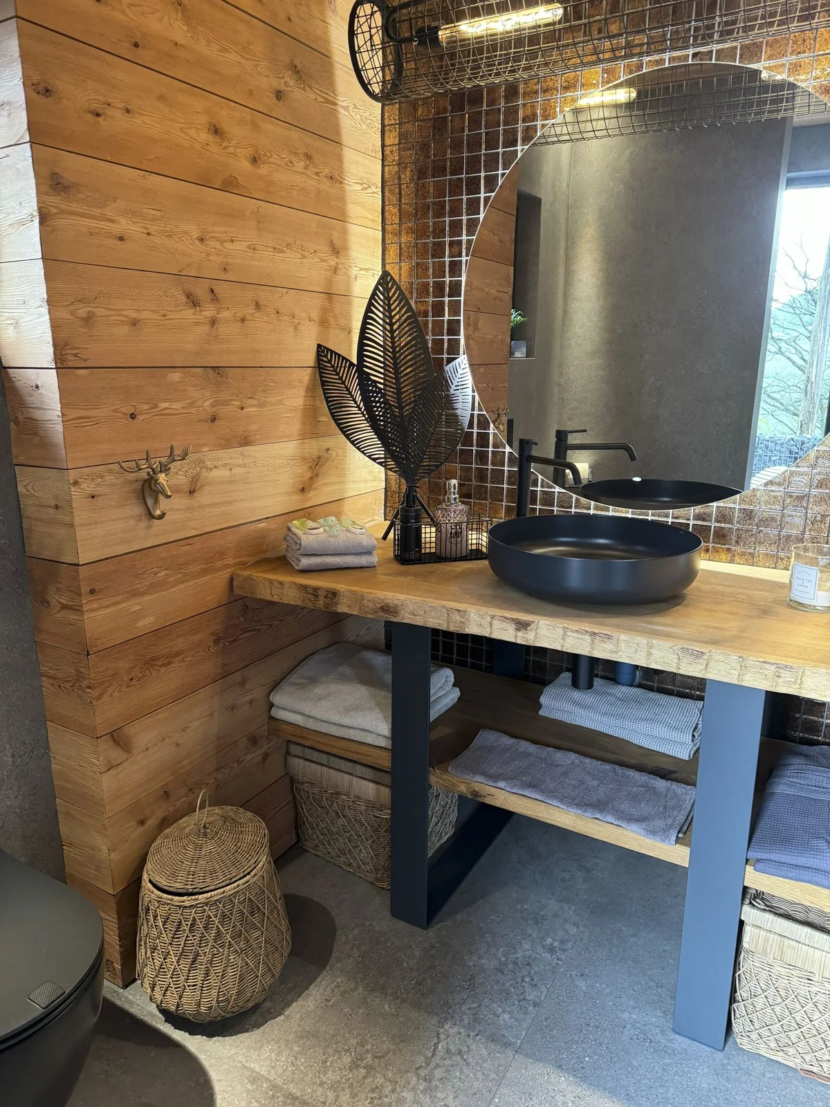Moderner Badezimmerbereich mit Holzwaschtisch, schwarzer runder Waschschüssel, großem Spiegel, Dekorationsgegenständen und Handtüchern, Wand im Backstein-Look, ruhige und natürliche Atmosphäre.