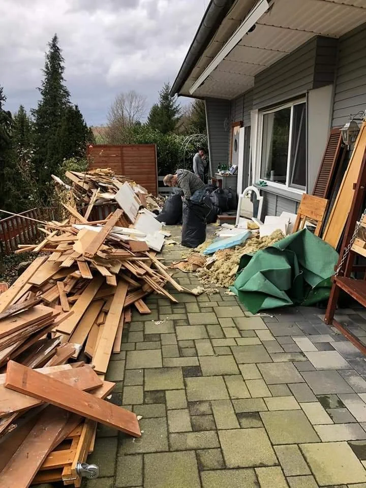 Auf einer Terrasse liegt eine große Menge an zerbrochenem Holz, Möbeln und Bauschutt. Zwei Menschen durchsuchen die Trümmer, während sich im Hintergrund ein Haus mit grauer Fassade befindet.