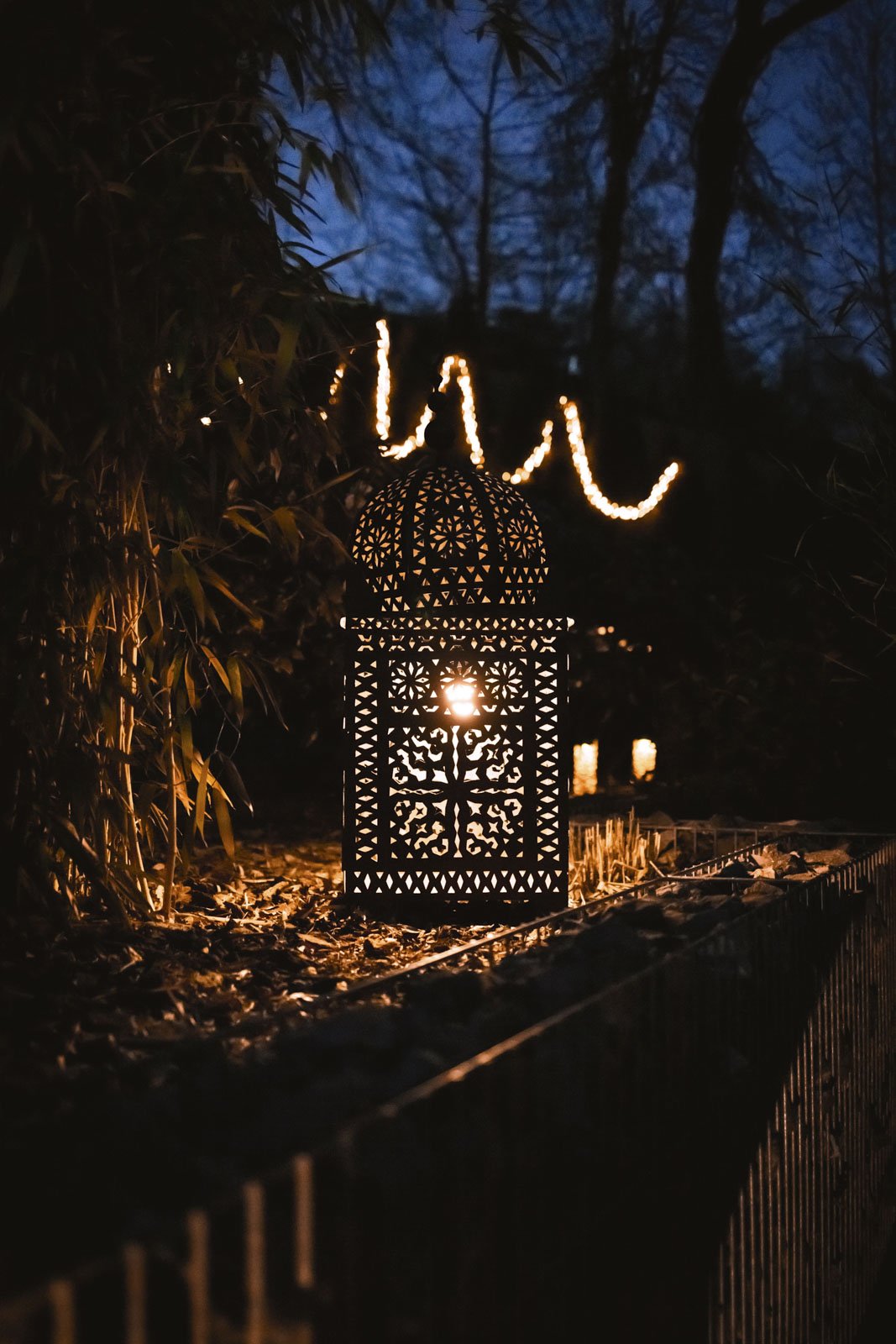 Eine mit Mustern verzierte chinesische Laterne, die im Dunkeln leuchtet, mit einer Lichterkette im Hintergrund und Bäumen gegen die blaue Dämmerung im Himmel.