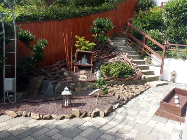 Ein terrassierter Garten mit Stufen, Pflanzen, Steinen, Dekorationen und einem roten Holzzaun.