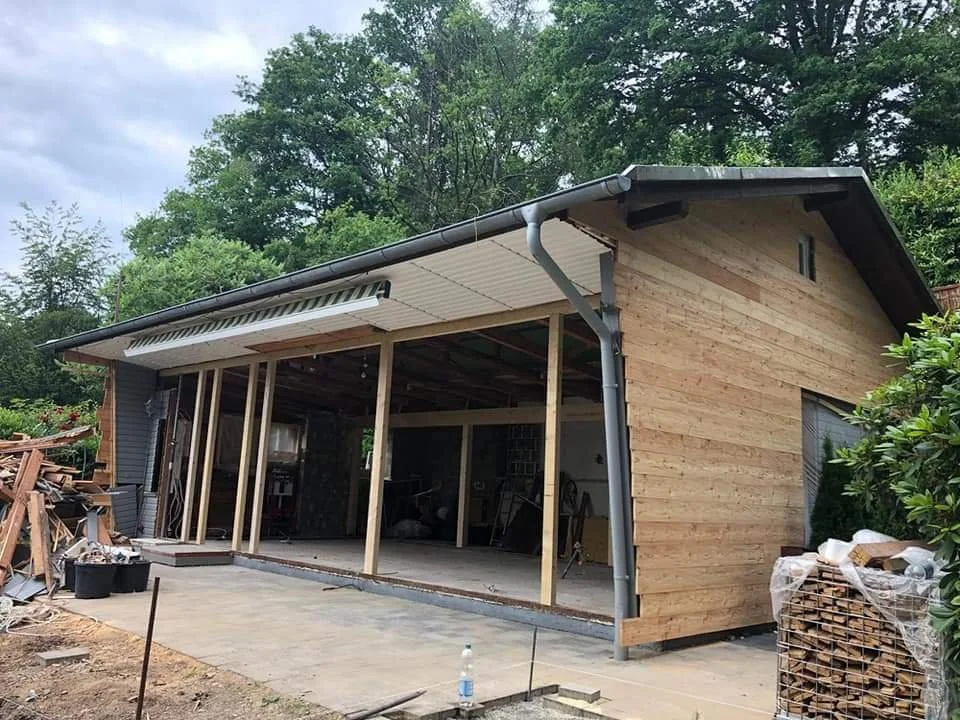 Ein Holzschuppen im Bau, mit Holzplanken verkleidet, offen an der Vorderseite, auf einem gepflasterten Boden, umgeben von Baum und Baustoffen.