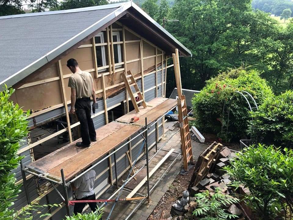 Ein Haus wird renoviert oder gebaut, mit Handwerkern auf einer temporären Plattform, um die Fassade und das Dach zu bearbeiten, umgeben von Bäumen und Gartenpflanzen.