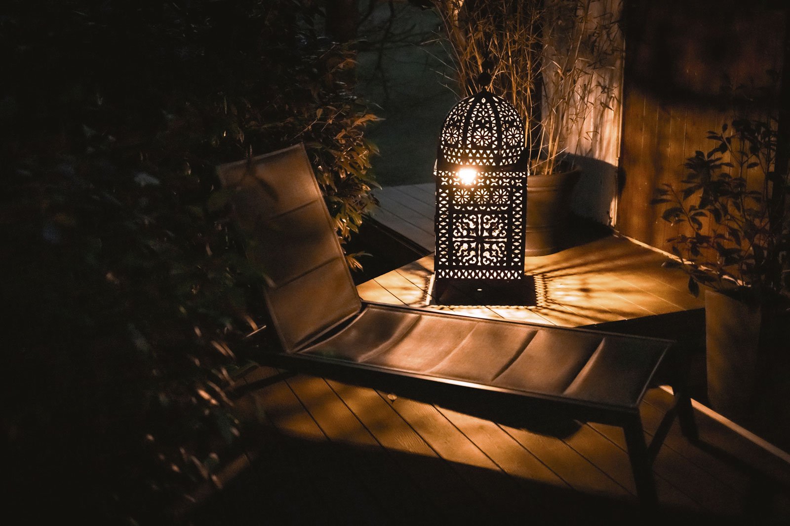 Schatten einer dekorativen Laterne auf einer Terrasse bei Nacht, umgeben von Pflanzen und einem Liegestuhl.