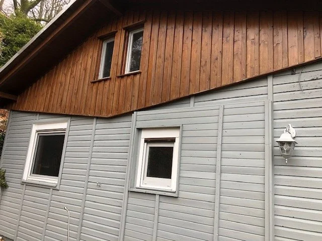Außenansicht eines Hauses mit grauem Holzverschalung und braunem Holz-Dachgeschoss, zwei weiße Fenster und eine Laterne an der Wand.