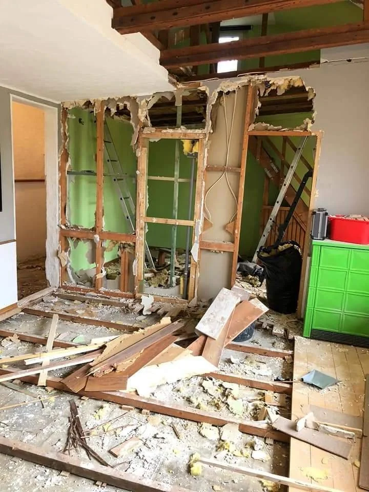 Der Innenraum eines Hauses in Renovierung mit demoliertem Wandabschnitt, herumliegender Holz- und Bauschutt, ein aufgestellter Leiter sowie eine grüne Möbelstück im rechten Bereich.