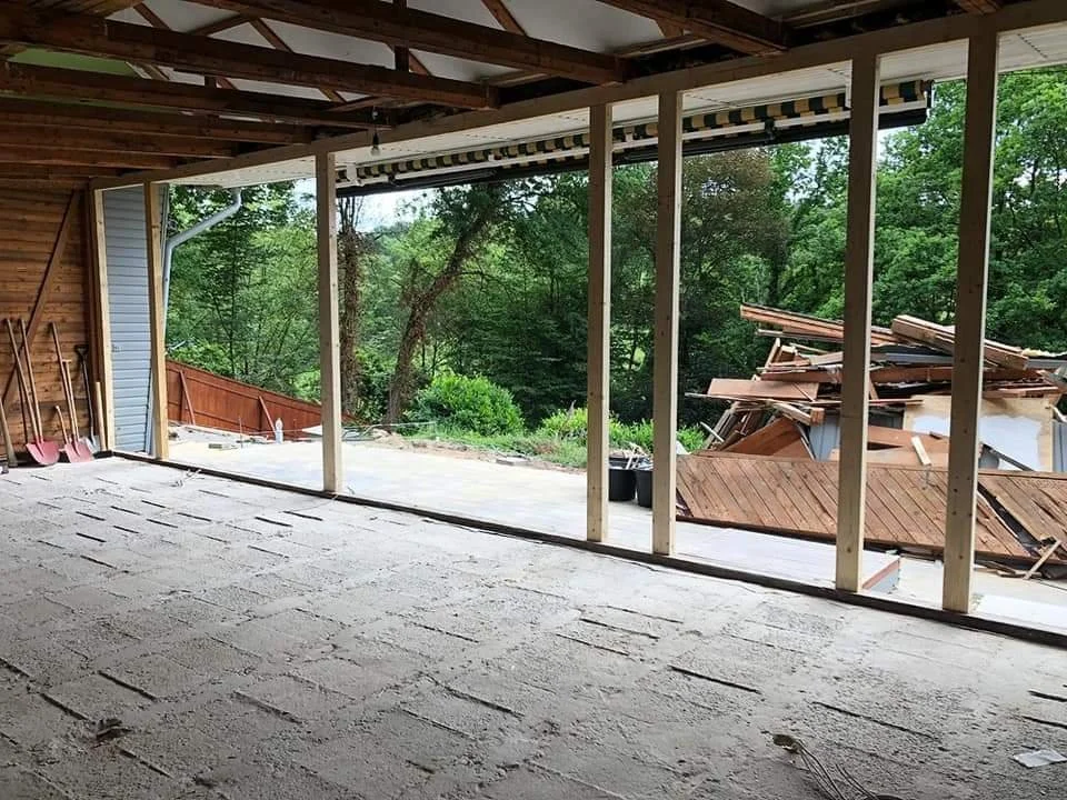 Baugerüst und Gerümpel vor einem Haus im Bau, Blick auf eine Terrasse mit Wald im Hintergrund.