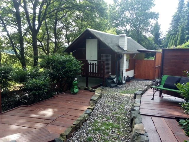 Garten mit Holzterrasse, kleiner Hüttenbereich, grünem Gartenzubehör, Bäumen und Zaunanlage im Hintergrund.