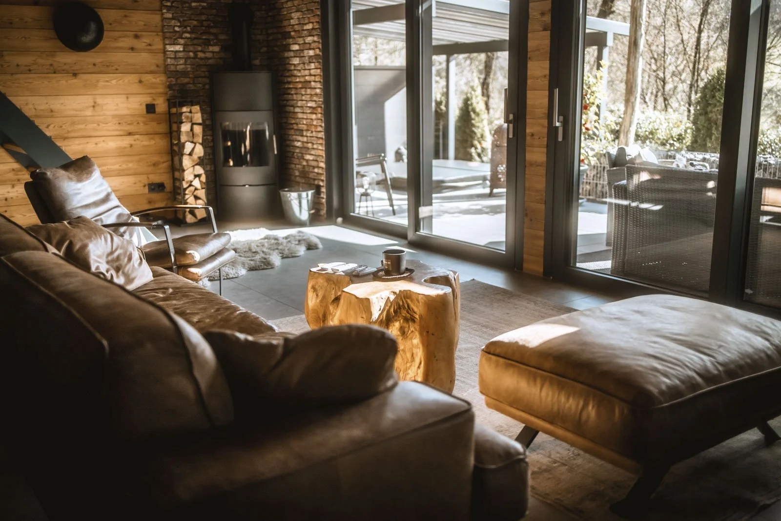 Gemütliches Wohnzimmer mit Leder-Couch, Holz-Tisch, Kamin, großen Glas-Schiebetüren und Terrasse mit Möbeln, Holz-Wandverkleidung, viel natürliches Licht.