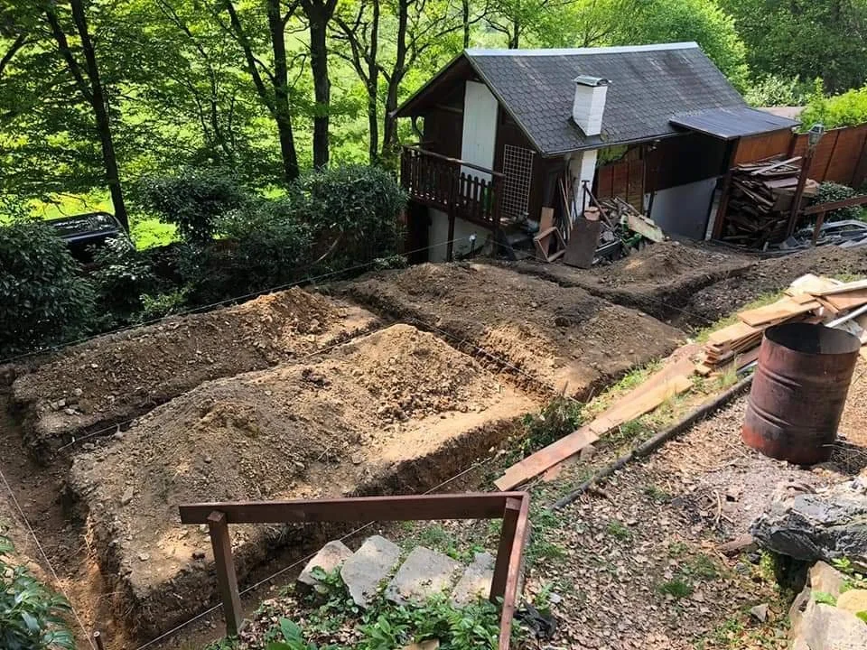 Baugrube im Garten neben einem kleinen Haus, mit Holz- und Eisenmaterialien sowie einem alten Metallfass.
