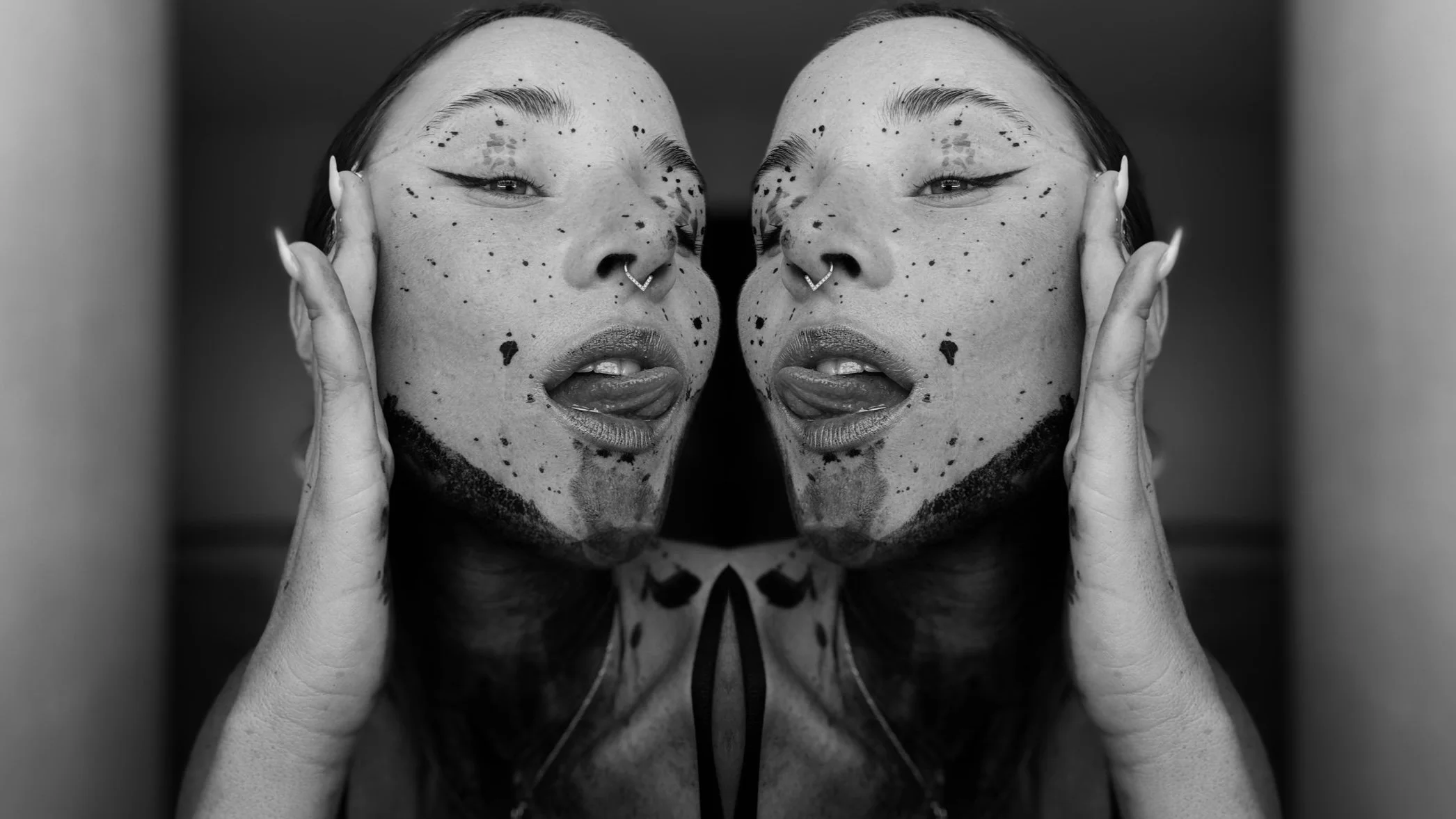 A symmetrical black-and-white photo of a person with tattoos, lipstick, piercing, and makeup, touching their face with both hands, with a mirror reflection effect.