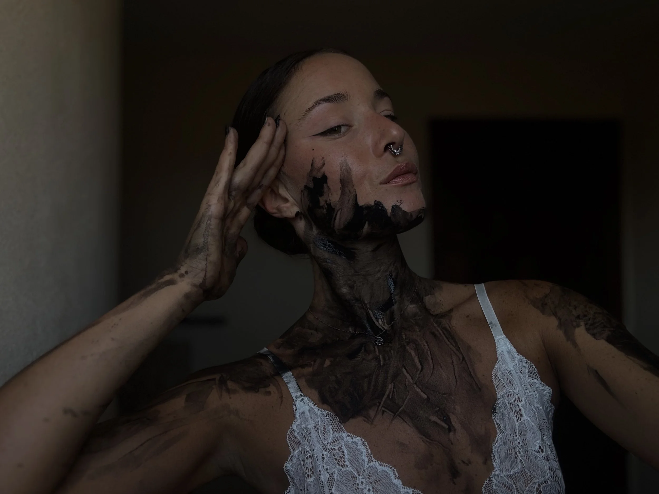 A woman with freckles and dark makeup on her face and arms, wearing lace lingerie, is being touched on her face by a hand covered in dark makeup or mud, against a dark background.