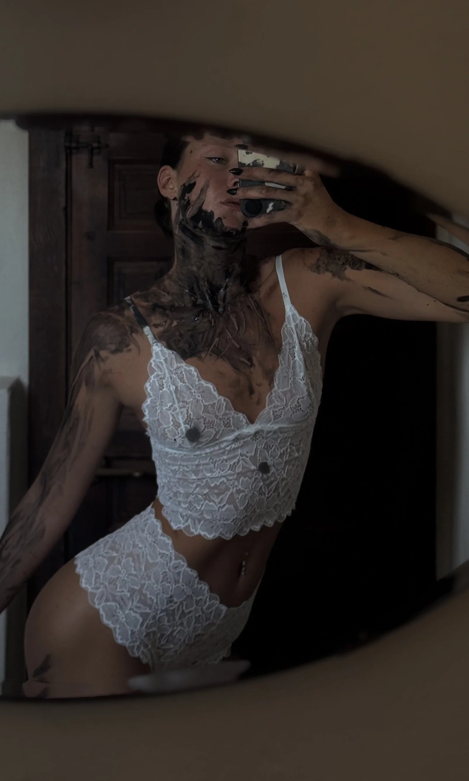 A woman taking a mirror selfie, wearing a white lace lingerie set, with black paint on her face and body. She has dark hair pulled back and is using a camera phone.