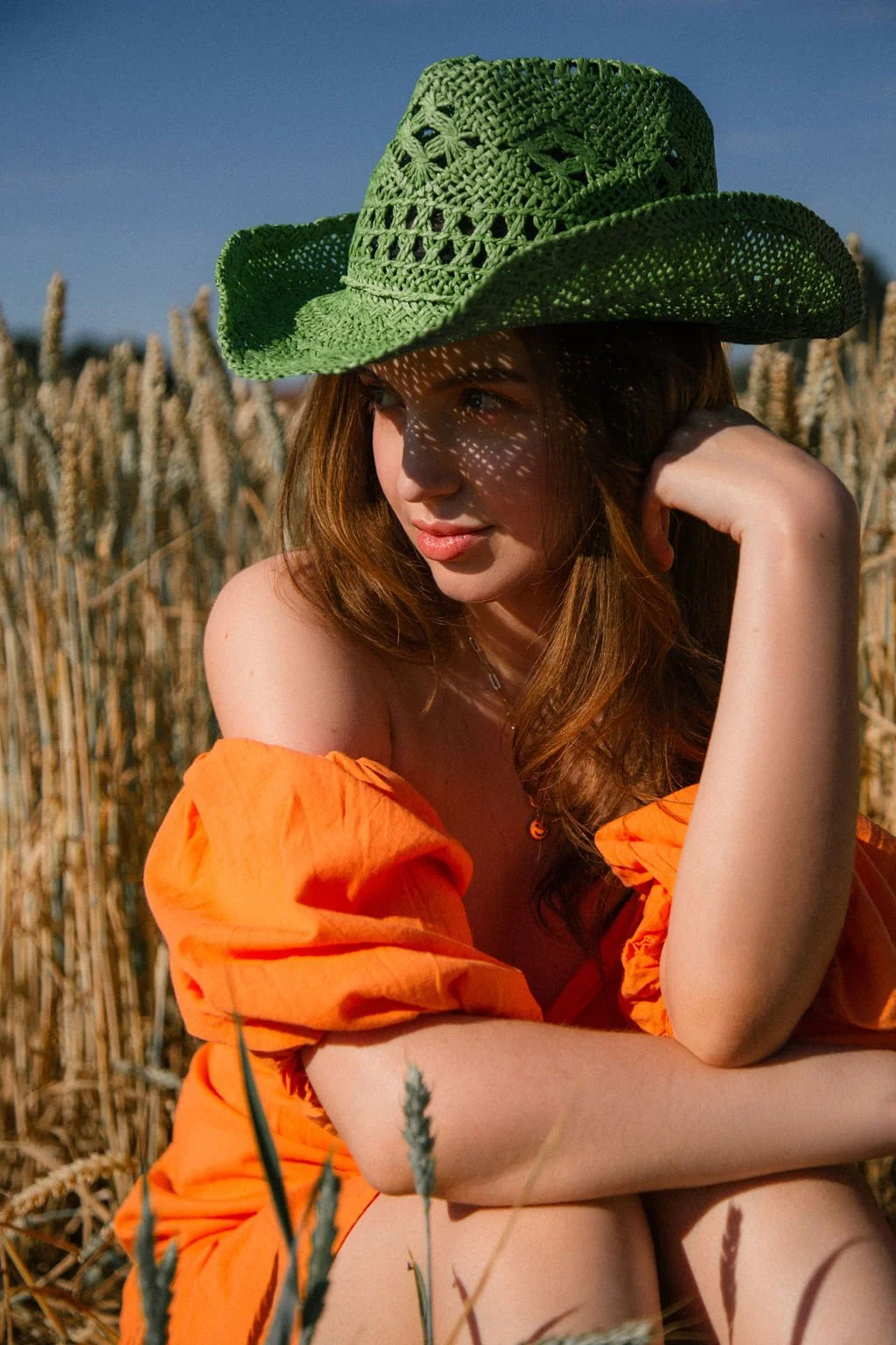 A woman with red hair wearing an orange off-shoulder top and a large green woven hat, sitting in a wheat field on a sunny day.