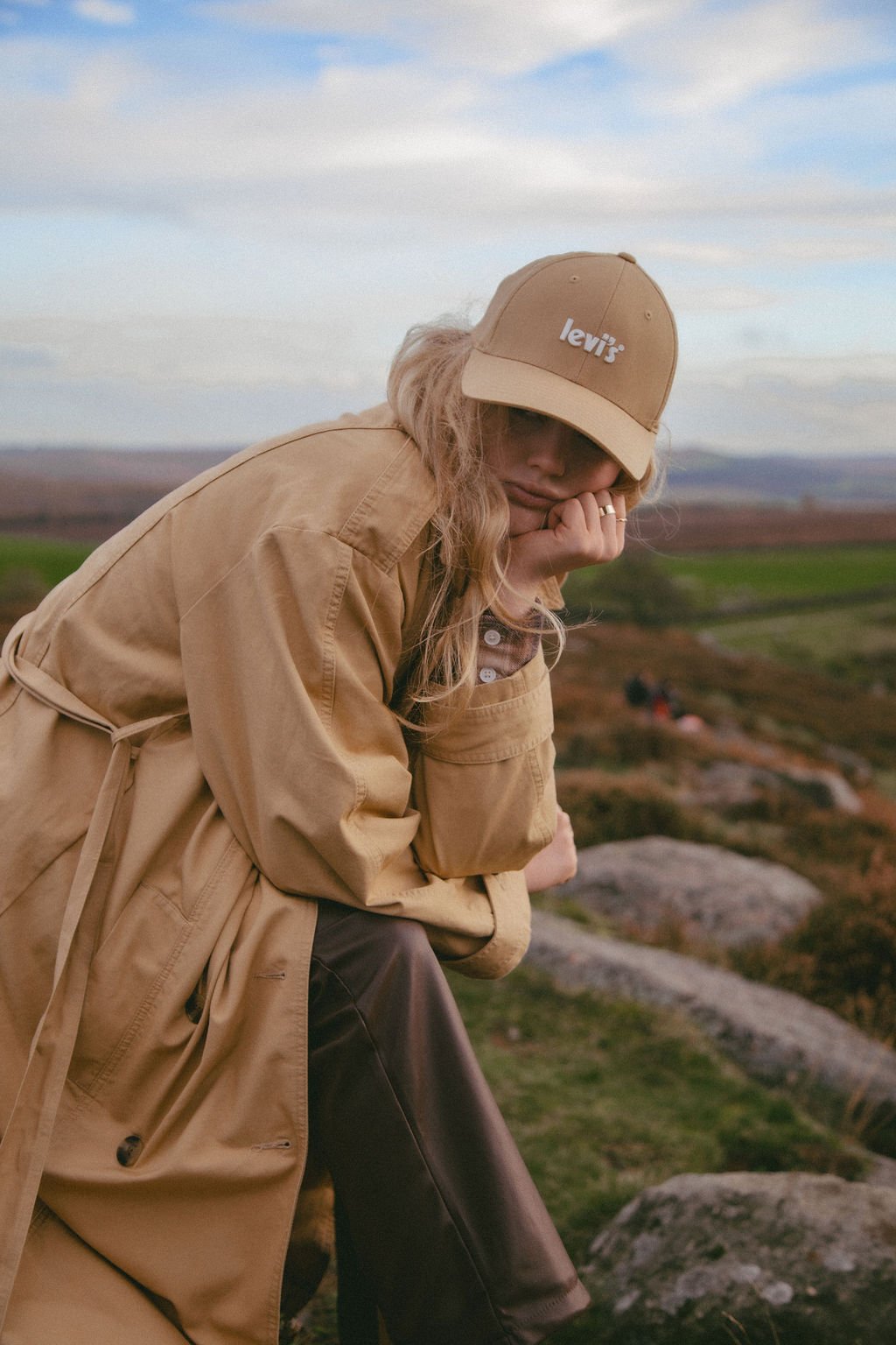A woman wearing a beige Levi's cap and trench coat, sitting outdoors on a rock, resting her chin on her hand with a scenic rural landscape background.