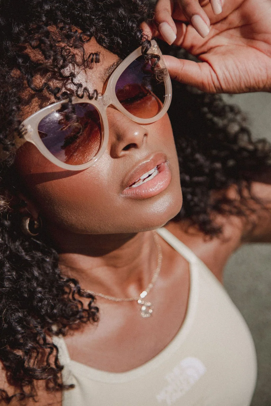 A woman with curly hair wearing beige sunglasses with reddish-brown lenses, a white tank top, and a delicate necklace, touching her sunglasses with her right hand.