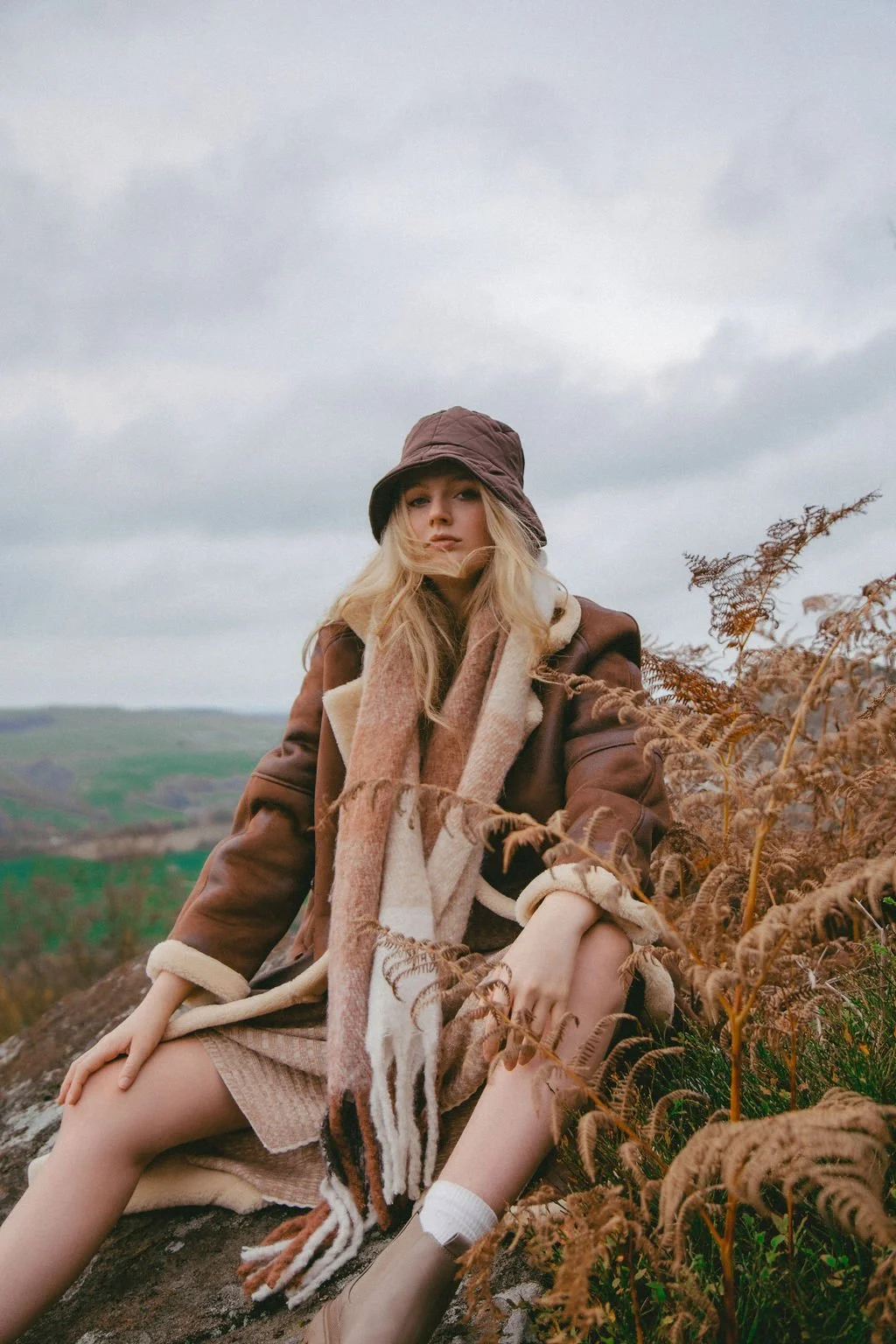 A young woman in a brown coat, beige dress, and a multicolored scarf sits on a rock outdoors, surrounded by dry ferns, with a cloudy sky and distant green landscape in the background.