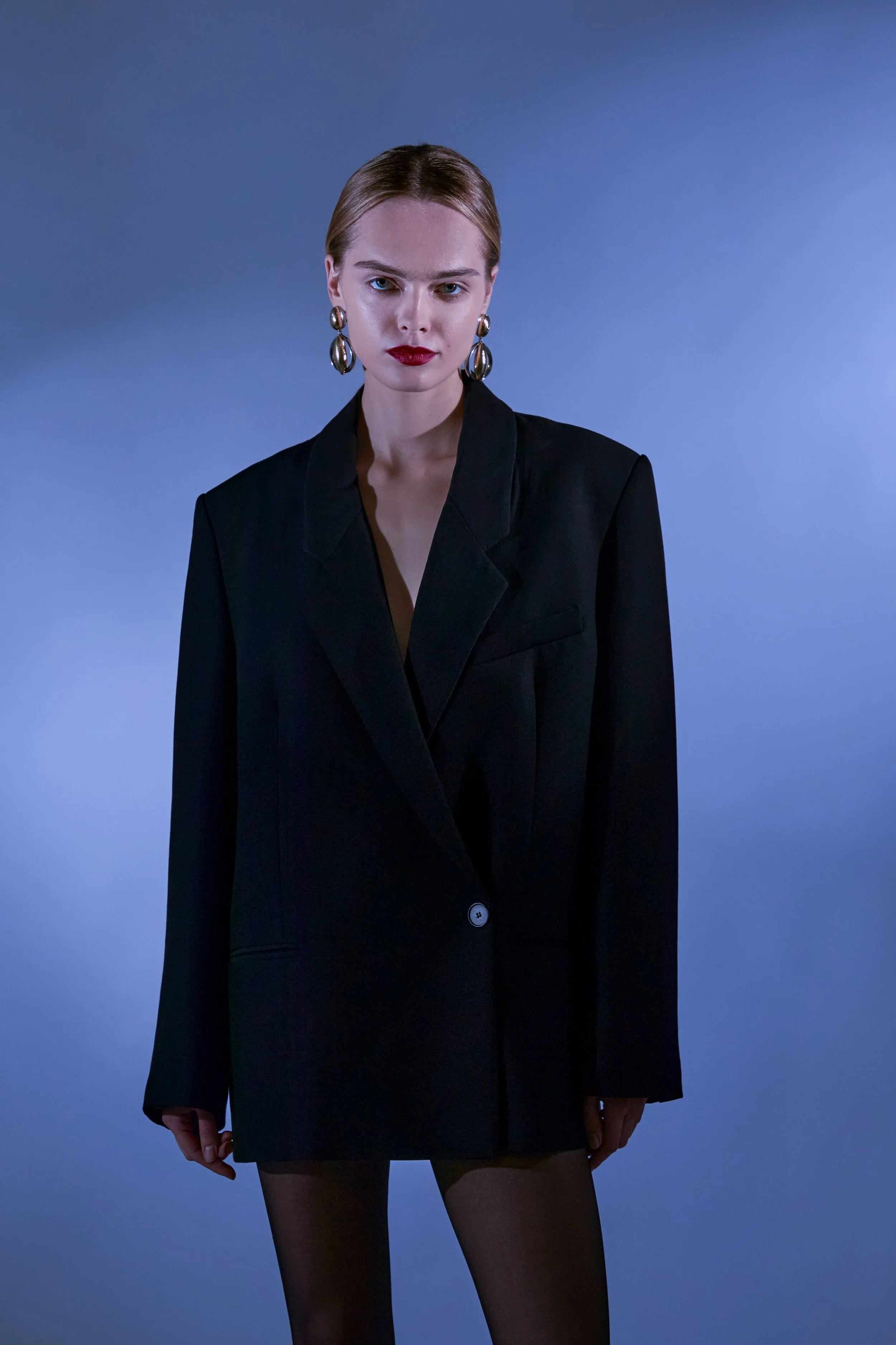 A woman with blonde hair pulled back, wearing a black blazer dress with a deep neckline, large silver earrings, red lipstick, standing against a blue gradient background.