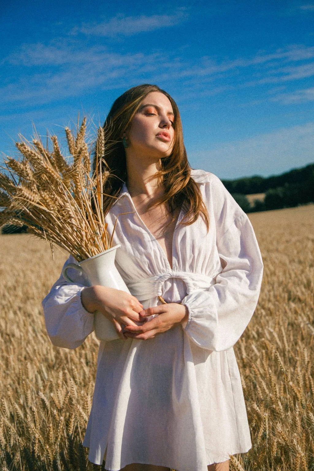 A woman with long brown hair standing in a wheat field, holding a vase with wheat stalks, with her eyes closed on a sunny day.