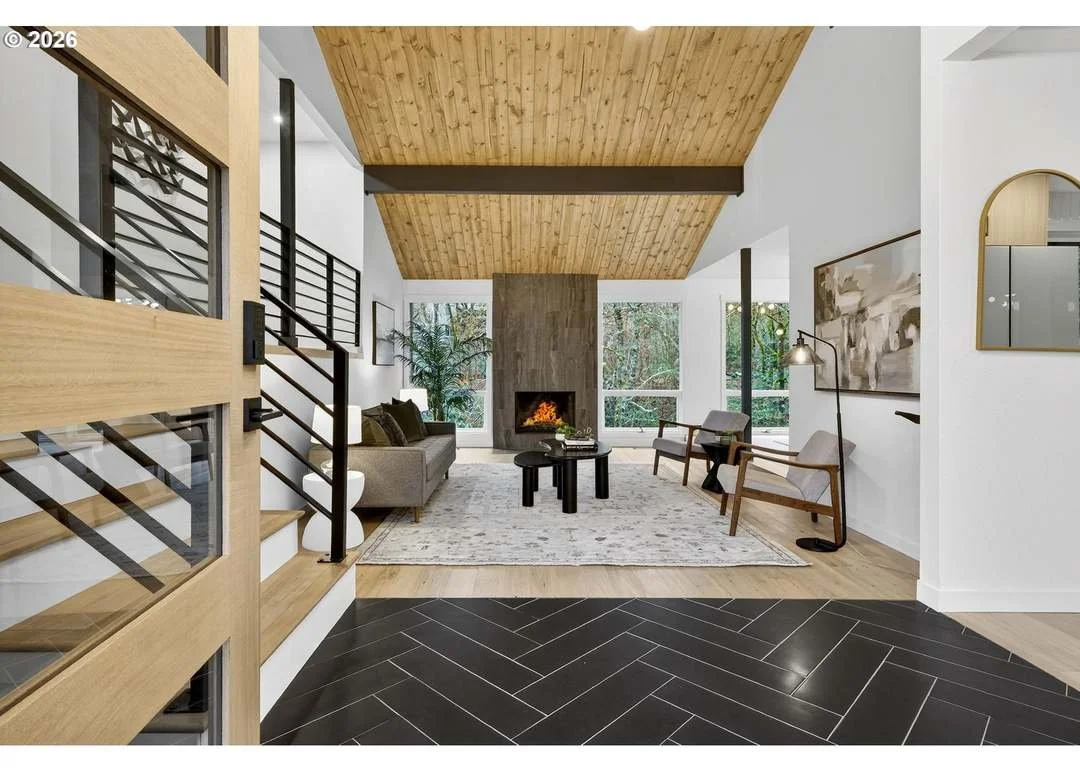 Modern living room with a wood-paneled ceiling, a fireplace, and large windows with a view of greenery outside.