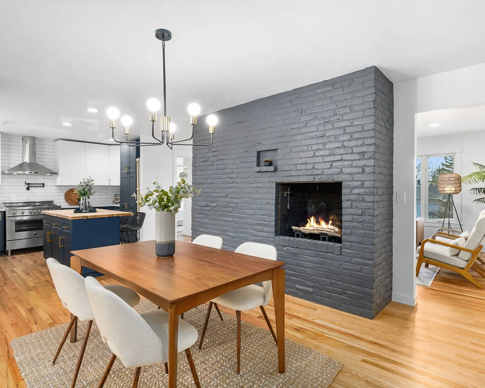 Open-concept living and kitchen area with a gray brick fireplace, modern chandelier, and wooden dining table with white chairs. The kitchen features white cabinets, a stainless steel stove, and a blue kitchen island. There is a vase with green foliage on the table, and to the right, a cozy sitting area with a chair, lamp, and large window showing trees outside.