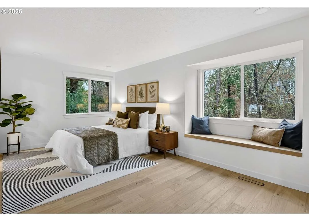 A minimalist bedroom with a white bed, brown and white bedding, two nightstands with lamps, a built-in window seat with cushions, large windows showing trees outside, and a potted plant in the corner.