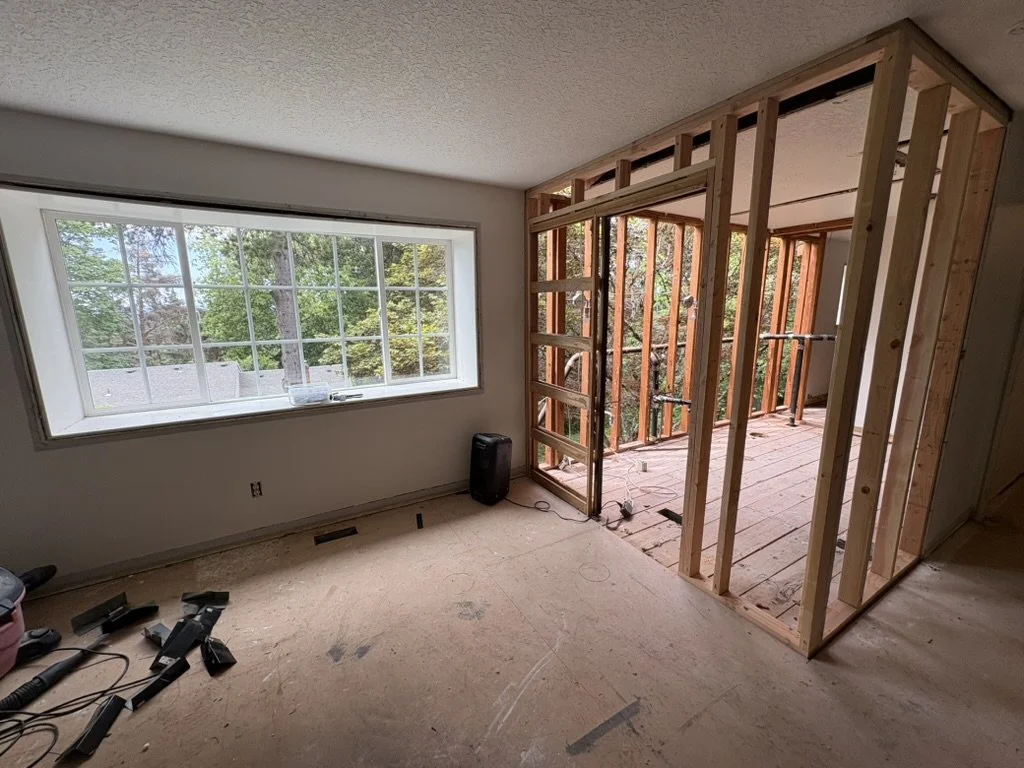 Room under renovation with exposed wooden framing for a new wall and a large window showing trees outside.