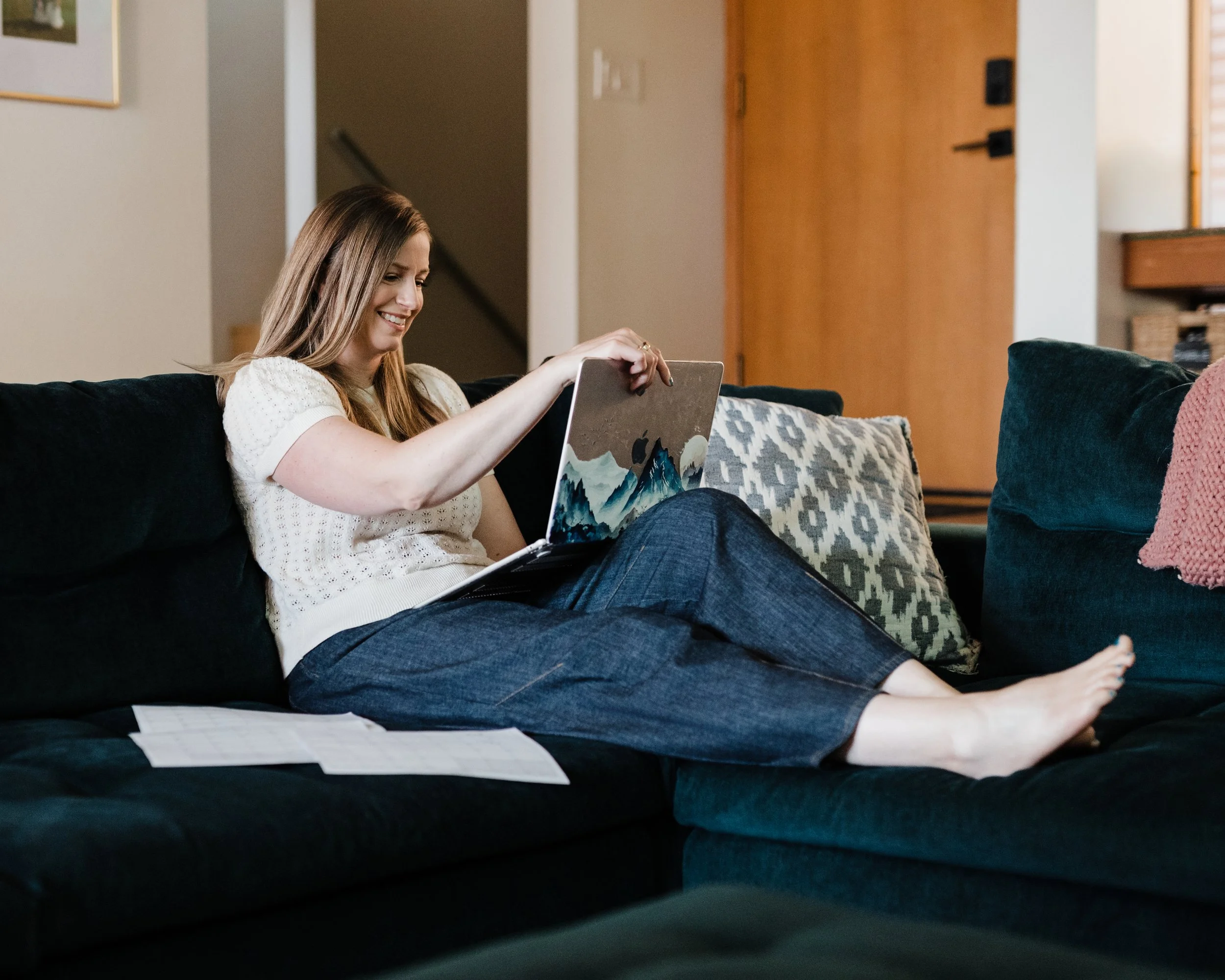 A woman with long, light brown hair sits on a dark green couch in a living room, smiling and looking at a tablet in her hand. She is wearing a white knit top and blue jeans. There are papers on her lap, and a pillow is next to her. The background shows a doorway, a staircase, and a wooden door.