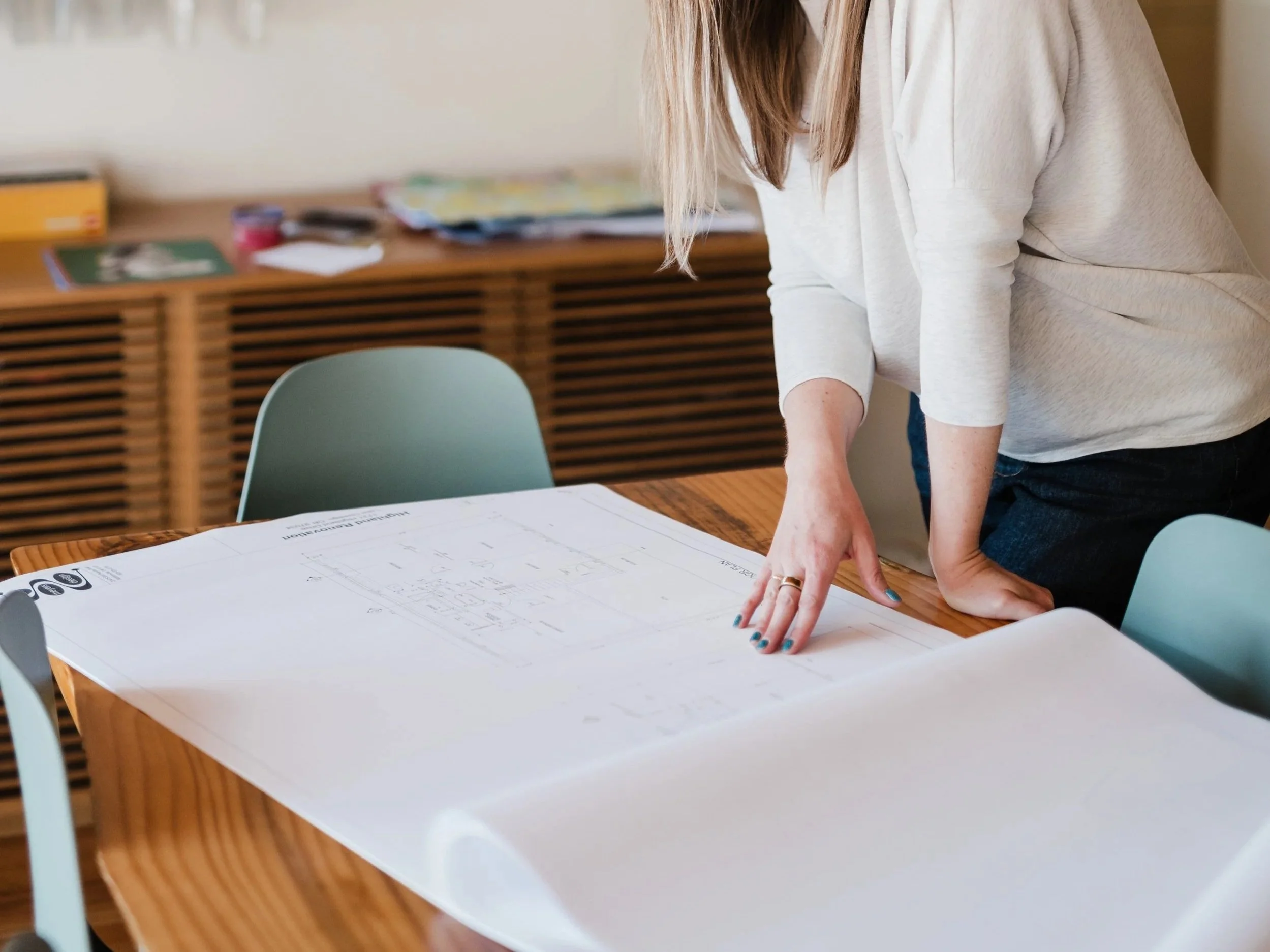 A person leaning over a table with architectural plans, wearing a light gray sweater, with stacked books and notebooks in the background.