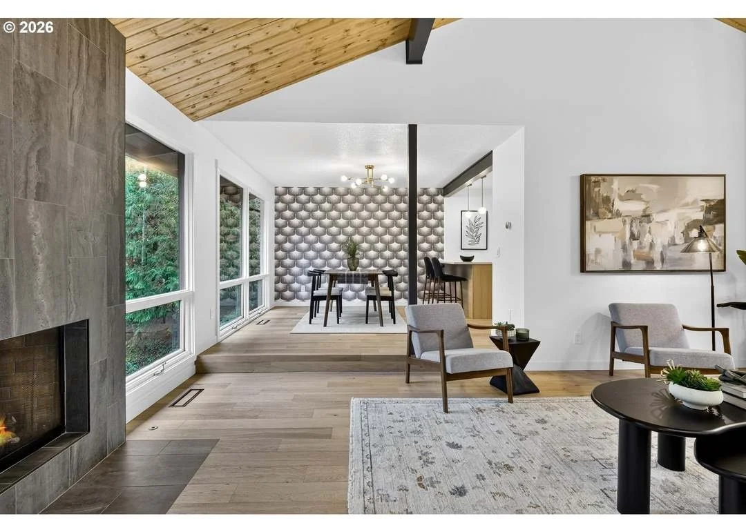 A modern living room with a fireplace, large windows, and a dining area in the background featuring a wall with a 3D geometric pattern.
