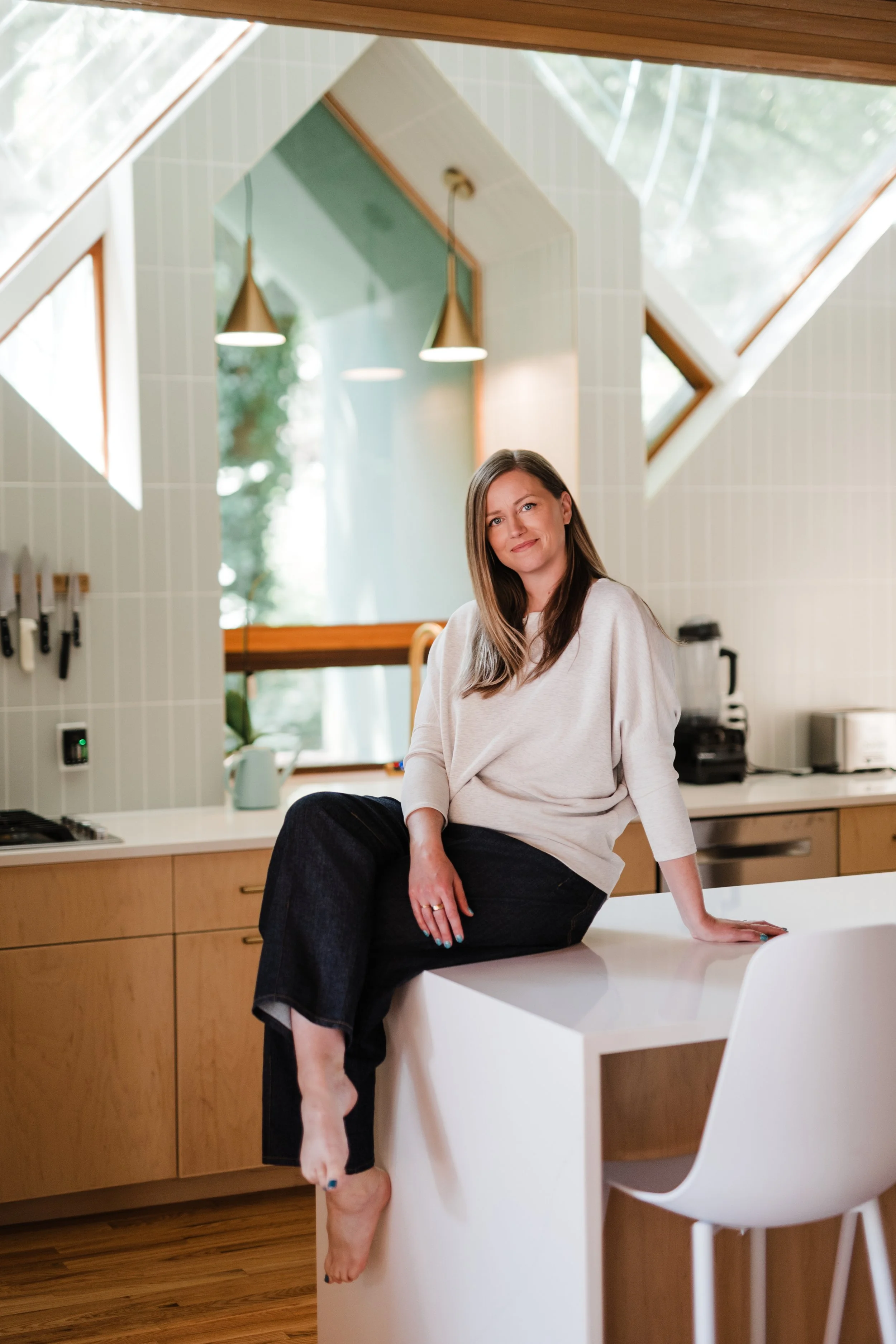 Woman sitting on a kitchen counter in a modern kitchen with large skylight windows, white appliances, and wooden cabinetry, smiling at the camera.