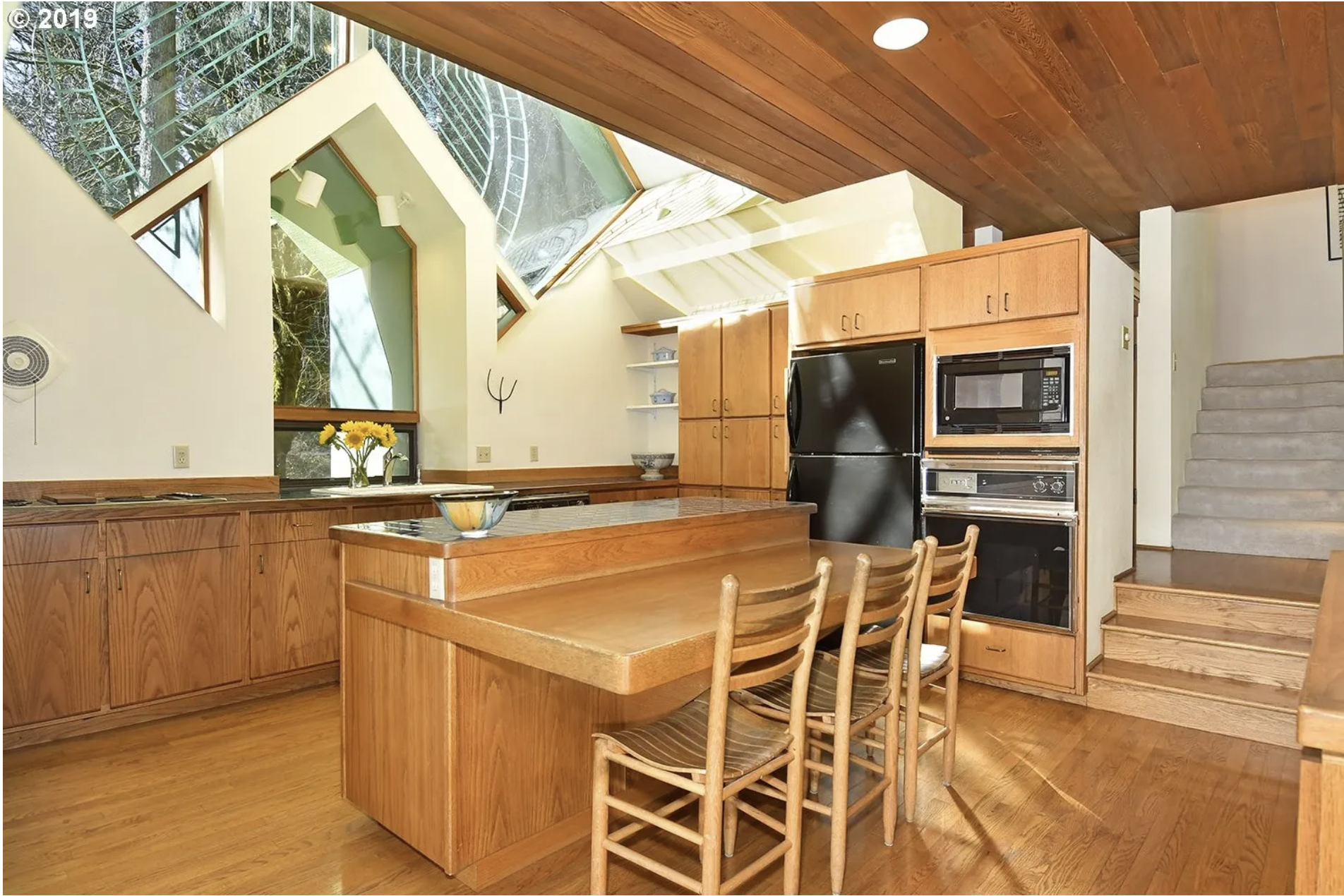 Interior view of a kitchen with wooden cabinetry, a kitchen island, a dining table with chairs, large angled windows with trees outside, and a wooden ceiling.