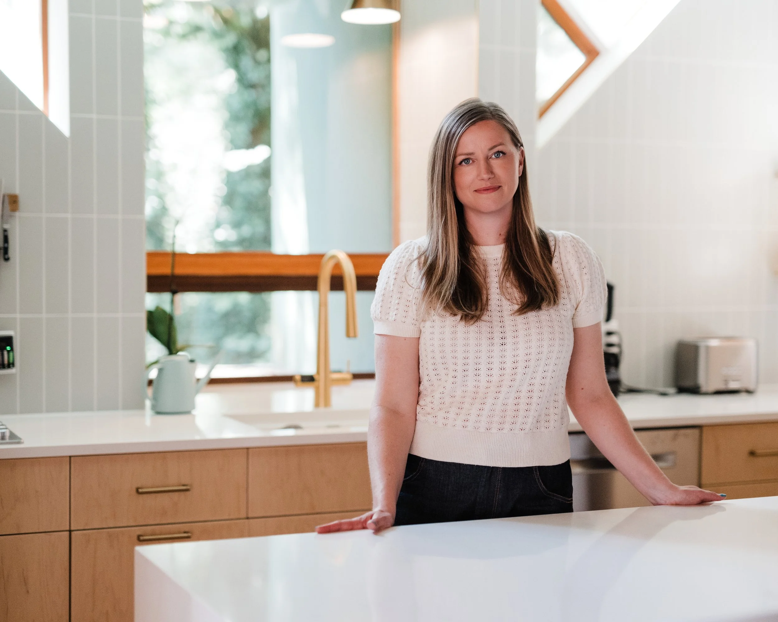 A woman with long hair stands in a modern kitchen, leaning on a white countertop with both hands, looking at the camera with a slight smile.
