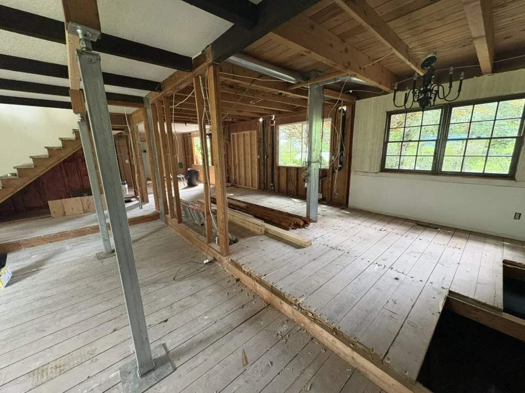 Interior of a house under renovation with exposed wooden framing, an unfinished floor, and a chandelier hanging from the ceiling. Windows reveal greenery outside.
